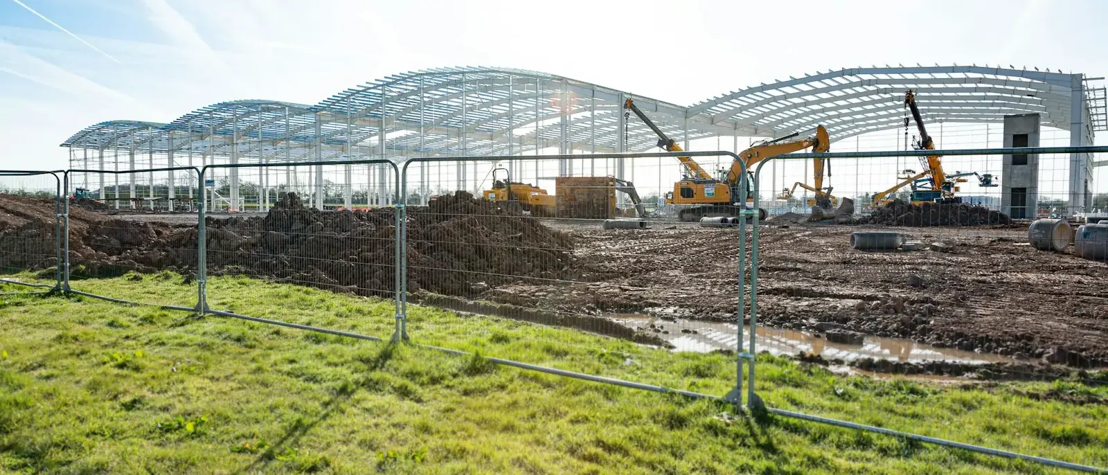 Pórticos de aço em arco e terças de um grande galpão em montagem, com escavadeiras no canteiro, etapa comum na construção de galpões em Goiânia