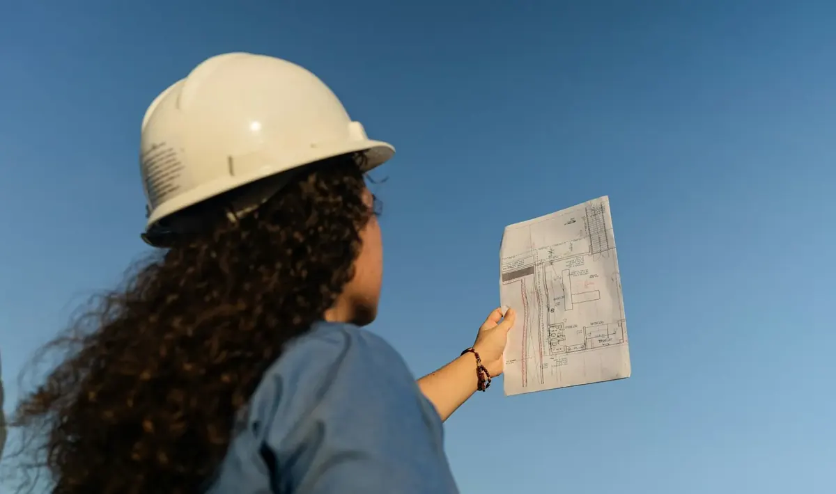 Profissional de capacete branco segurando uma planta impressa contra o céu, conferência típica do gerenciamento de obras em Goiânia