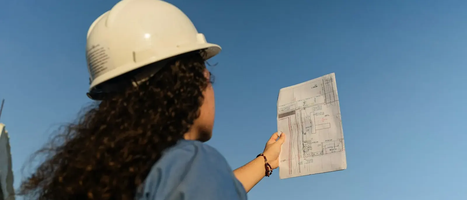 Profissional de capacete branco segurando uma planta impressa contra o céu, conferência típica do gerenciamento de obras em Goiânia