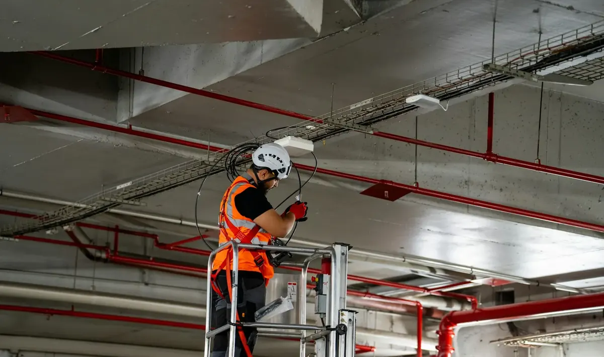 Técnico em plataforma elevatória ajustando cabos junto ao teto de uma garagem com tubulação vermelha, rotina de manutenção predial em Goiânia