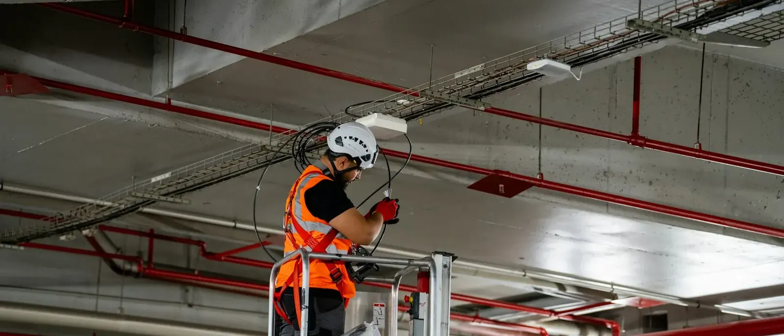 Técnico em plataforma elevatória ajustando cabos junto ao teto de uma garagem com tubulação vermelha, rotina de manutenção predial em Goiânia
